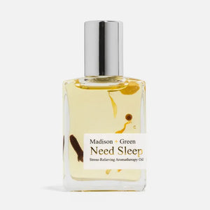 A clear glass bottle labeled “MADISON AND GREEN - NEED SLEEP AROMATHERAPY BODY OIL MINI” by MADISON GREEN, filled with yellow botanical body oil and visible botanicals, topped with a silver cap and photographed against a plain white background. Crafted to promote relaxation and tranquility, this calming oil delivers soothing aroma with every use.