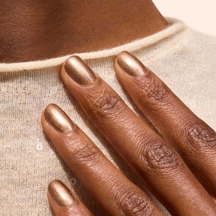 A hand with shimmery bronze vegan, cruelty-free nail polish holds a matching bottle labeled “MANUCURIST - GREEN™ Nail Polish in Tanned” against a brown sweater.