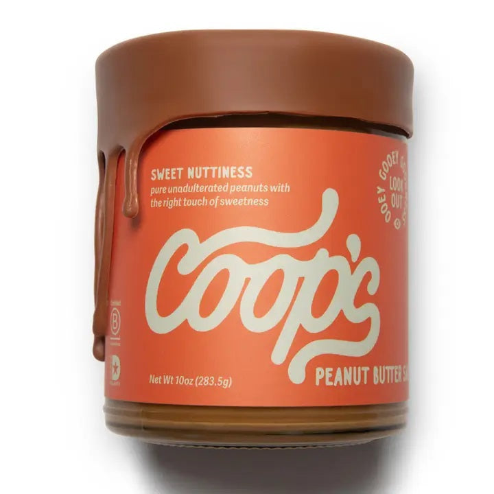 A 10 oz (283.5 g) jar of COOPS Peanut Butter Sauce, handmade in small batches to perfection, features chocolate-like drips down the lid and an orange label with “Sweet Nuttiness” and “Peanut Butter Sauce.”.