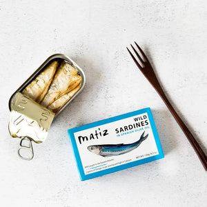A blue rectangular box of Matiz España Wild Caught Smoked Sardines in organic olive oil, with a dash of sea salt. The illustration highlights the product's high Omega-3 and calcium content. Net weight: 120g (4.2 oz). These wild-caught sardines hail from the Eastern Atlantic for an authentic flavor experience.