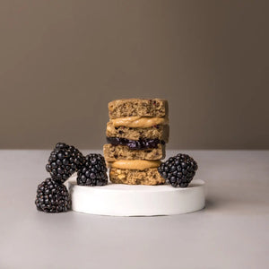 A purple STARS+HONEY - PEANUT BUTTER BLACKBERRY protein bar rests on a beige surface, with its wrapper highlighting 10g collagen, 15g protein, and 2g sugar.