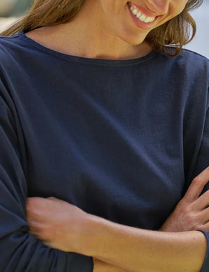 A woman with wavy brown hair stands outdoors among greenery and rocks, facing away but looking over her shoulder with a smile. She wears the FRANK & EILEEN Ella Long Sleeve Boatneck Tee in Classic Navy, paired with white pants.
