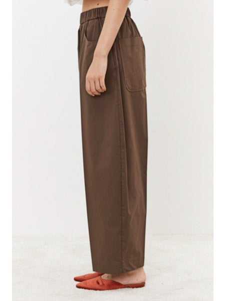 A person is seen from behind wearing MEIMEIJ&#39;s Pull On Cotton Pant, featuring an elastic waistband for comfort. The loose-fitting brown pants with large back pockets are paired with a white top peeking out at the waist, standing on a light surface.
