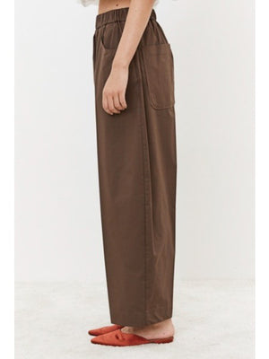 A person is seen from behind wearing MEIMEIJ's Pull On Cotton Pant, featuring an elastic waistband for comfort. The loose-fitting brown pants with large back pockets are paired with a white top peeking out at the waist, standing on a light surface.
