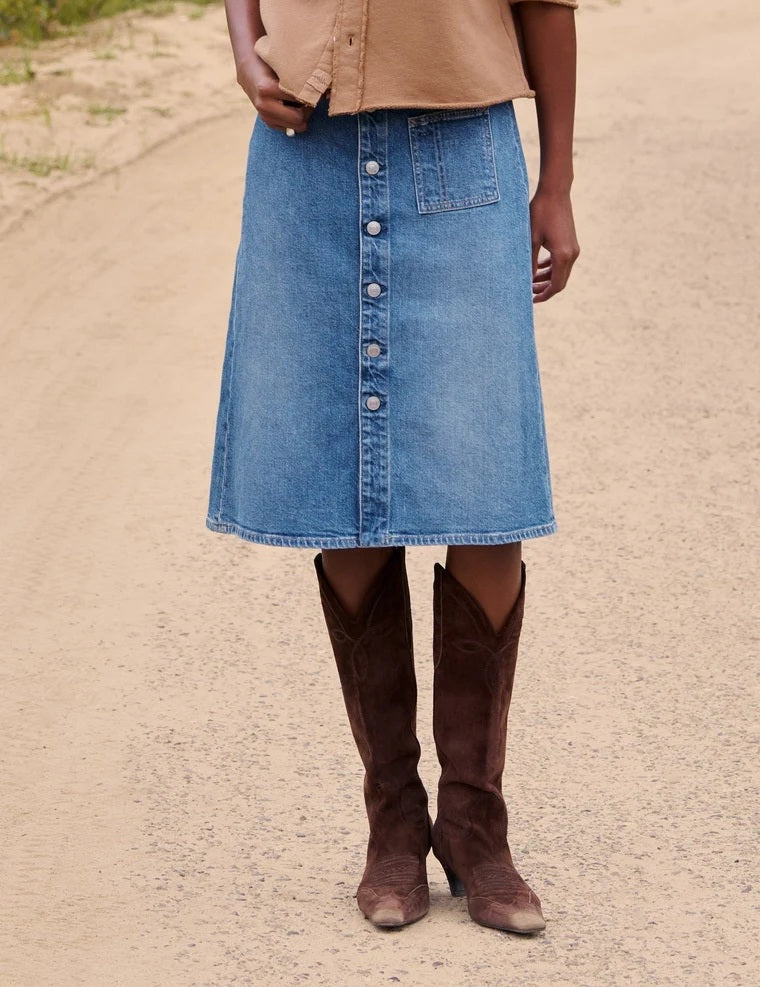 A woman walks on a dirt road near a fence, wearing a tan button-up shirt and the FRANK & EILEEN - SHANNON CLASSIC DENIM SKIRT IN 1978 WASH, paired with brown boots. In the background, trees and horses stand in a grassy field.