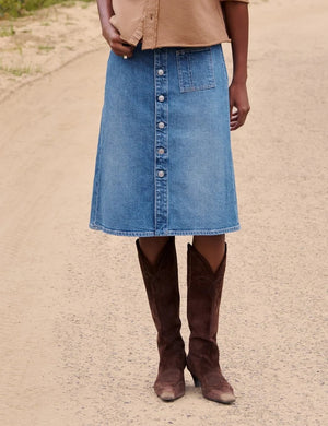 A woman walks on a dirt road near a fence, wearing a tan button-up shirt and the FRANK & EILEEN - SHANNON CLASSIC DENIM SKIRT IN 1978 WASH, paired with brown boots. In the background, trees and horses stand in a grassy field.