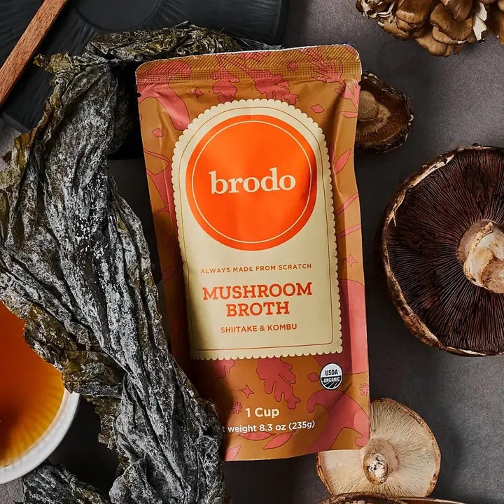 BRODO BROTH COMPANY’s BRODO BROTH- MUSHROOM ORGANIC pouch sits on a table amidst dried seaweed, whole mushrooms, and a bowl of broth, creating an earthy, natural scene that emphasizes this keto-friendly and dairy-free option.