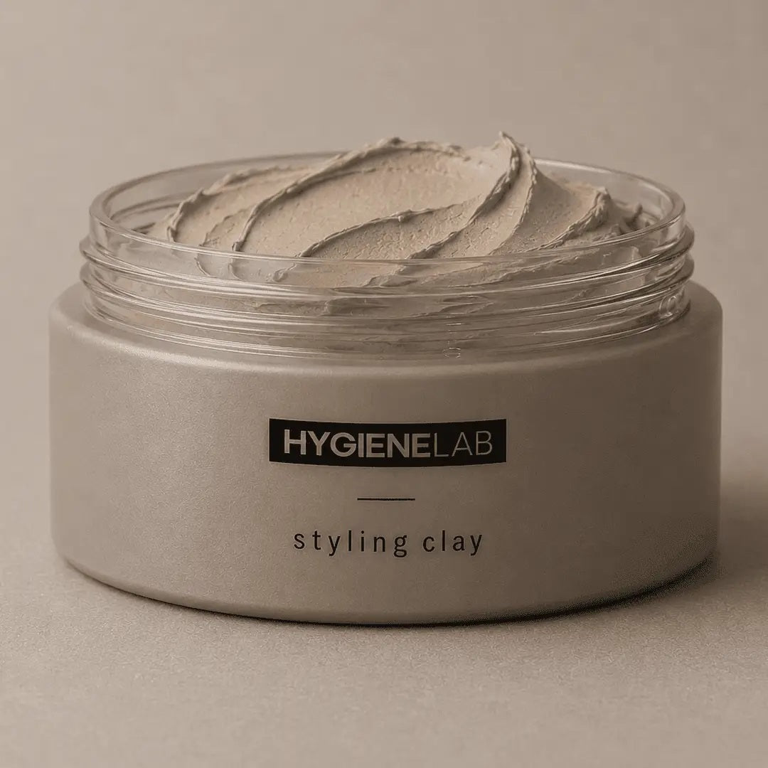 An open, round container of HYGIENELAB - STYLING CLAY displays the smooth, light gray clay inside with a swirled surface. This styling clay from HYGIENELAB provides a medium-strong hold and a natural matte finish.