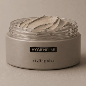 An open, round container of HYGIENELAB - STYLING CLAY displays the smooth, light gray clay inside with a swirled surface. This styling clay from HYGIENELAB provides a medium-strong hold and a natural matte finish.