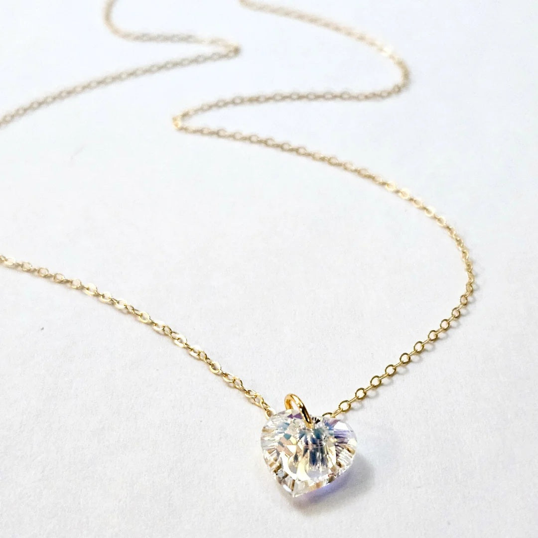 The VERDIER - AURORA CRYSTAL HEART NECKLACE from VERDIER features a sparkling crystal AB heart-shaped pendant suspended on a delicate 14k gold filled chain, shimmering against a white background.