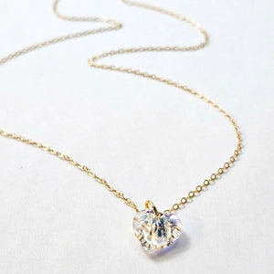 The VERDIER - AURORA CRYSTAL HEART NECKLACE from VERDIER features a sparkling crystal AB heart-shaped pendant suspended on a delicate 14k gold filled chain, shimmering against a white background.