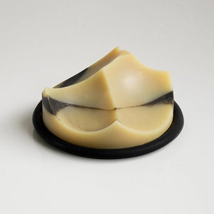 The UME STUDIO UME - CEDAR SANDALWOOD ERODE SOAP, featuring curved geometric shapes in beige and black, rests on a round black dish against a white background, showcasing its botanical formula with essential oils.