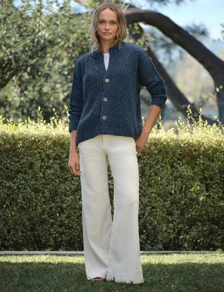 A person with wavy blonde hair, wearing a relaxed fit FRANK & EILEEN Montecito Crew Neck Cardigan in Pure Italian Cotton Blueberry and white pants, stands outdoors facing away, surrounded by greenery and trees.