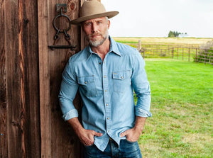 A man with a gray beard wears the CRIQUET WESTERN DENIM SHIRT WITH A PEARL SNAP IN LIGHT WASH by CRIQUET as he stands outdoors near a metal fence, with green grass and an open field in the background.