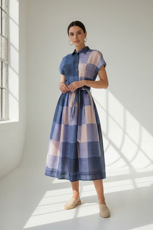 A woman wearing the LAVI - BELLELL Dress in Plaid by LAVI, a short-sleeve, collared shirt dress with a blue and beige plaid pattern and a tied waist, accessorized with gold earrings, bracelets, and open-toed heels, stands against a plain white background.