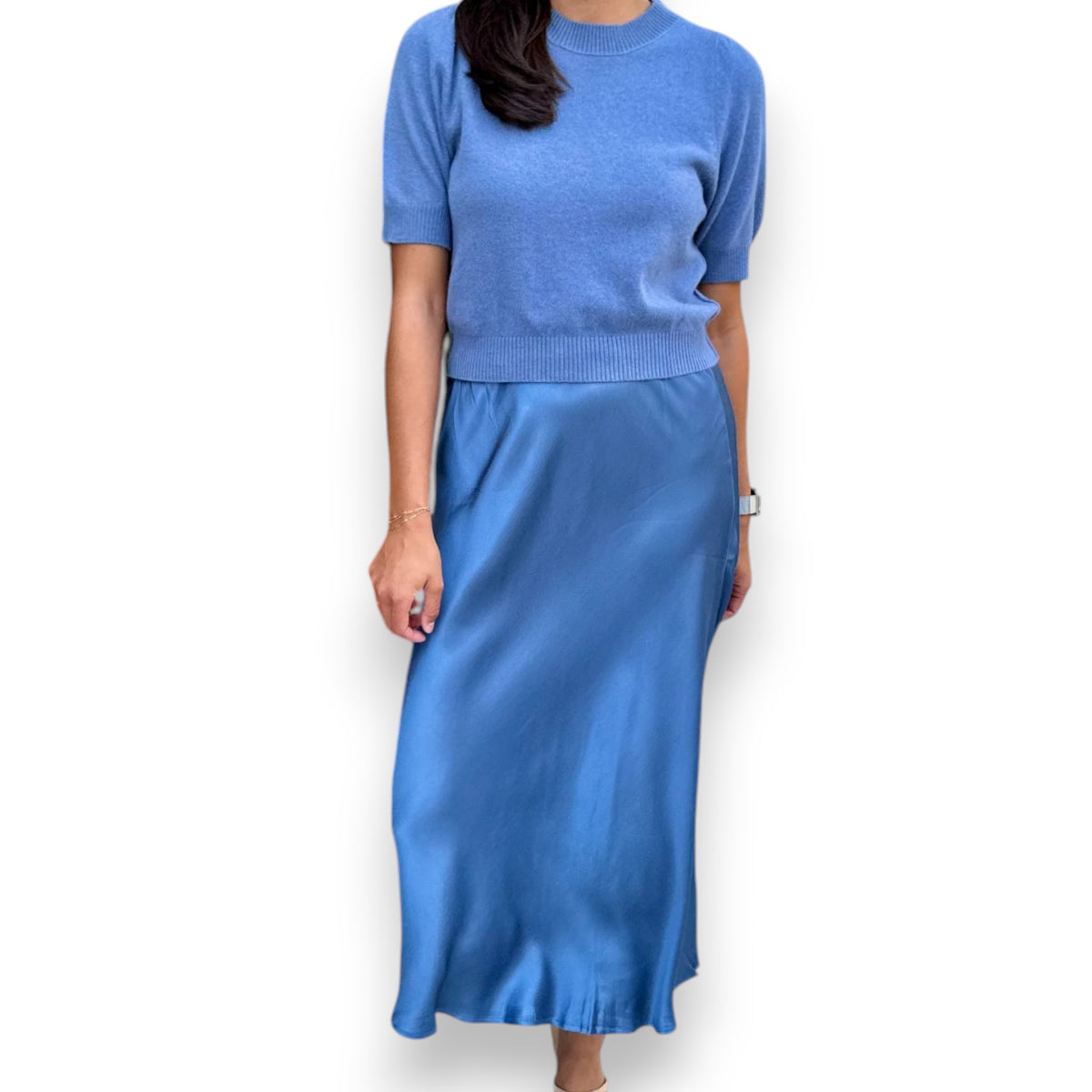 A woman models the ROSE &amp; CROWN - SATIN MIDI SKIRT in silky blue, paired with a short-sleeved light blue knit top, standing against a white background. Her head is not visible in the image.