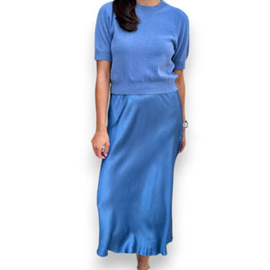 A woman models the ROSE & CROWN - SATIN MIDI SKIRT in silky blue, paired with a short-sleeved light blue knit top, standing against a white background. Her head is not visible in the image.