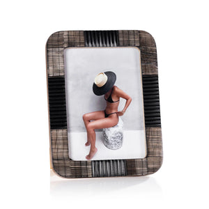 Two ZODAX MAJORCA BLACK CHISELED HORN 5x7 photo frames showcase images of a woman—one in a black swimsuit holding a drink, the other seated on rocks in a sunhat. A dark decorative piece accents the resin frames in the background.