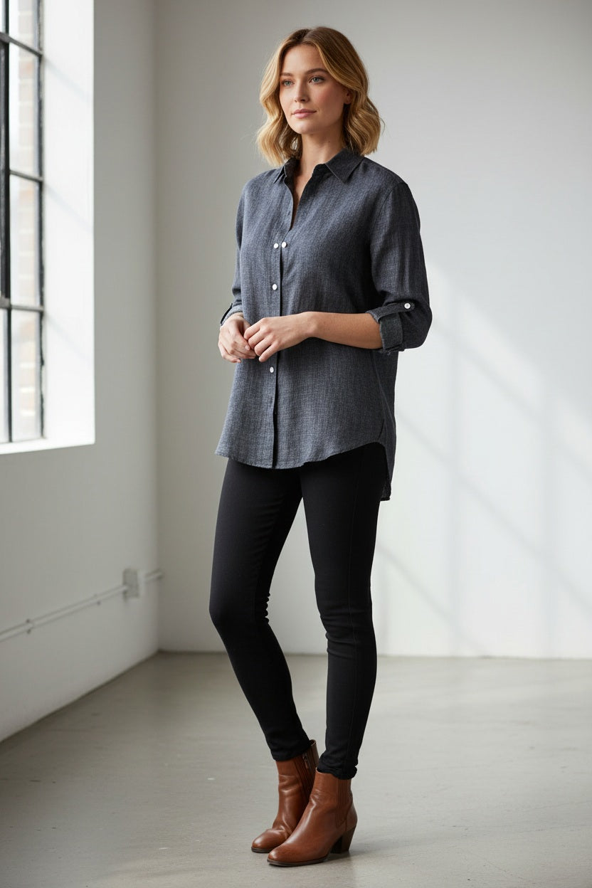 A person with short brown hair, dressed in the EMILY PHILLIPS - THE LINEN DENIM SHIRT—a long-sleeve, button-up gray shirt from EMILY PHILLIPS crafted from breathable linen fabric—and black pants stands against a plain light background, looking slightly to the left with a neutral expression.