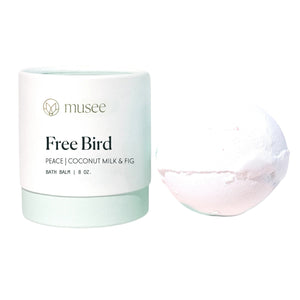 A light green and white container labeled “MUSEE - FREE BIRD THERAPY BATH BALM” sits beside a white bath bomb, offering a soothing and therapeutic bath experience with natural ingredients and coconut essential oil.
