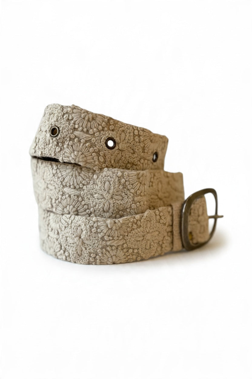 The JENNY KRAUSS - DESSERT DREAMS BELT by JENNY KRAUSS is a beige, hand-embroidered Peruvian belt with intricate floral motifs, metal eyelets, and a silver buckle, artfully coiled on a light surface.
