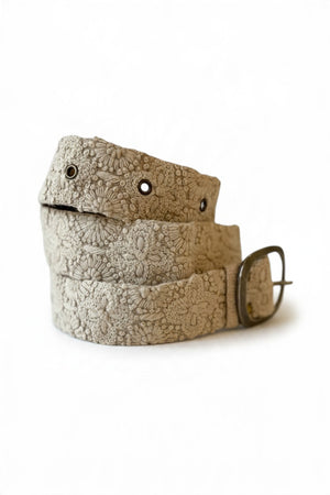 The JENNY KRAUSS - DESSERT DREAMS BELT by JENNY KRAUSS is a beige, hand-embroidered Peruvian belt with intricate floral motifs, metal eyelets, and a silver buckle, artfully coiled on a light surface.