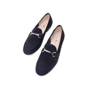 The MICHELE LOPRIORE - LEYLA LOAFER is a navy blue suede loafer adorned with a silver horsebit ornament on the vamp. Made in Italy and crafted from genuine leather, this shoe features a low heel and boasts a sleek, streamlined silhouette against a plain, off-white textured surface.