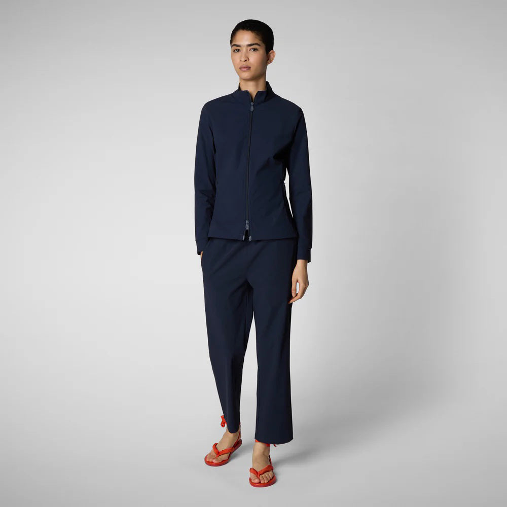 A person with shoulder-length hair and a neutral expression wears a white long-sleeve shirt, SAVE THE DUCK MILAN PULL ON PANT in black wrinkle-free fabric, and black slip-on shoes. Their hands rest at their sides against a plain background.