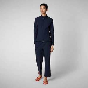 A person with shoulder-length hair and a neutral expression wears a white long-sleeve shirt, SAVE THE DUCK MILAN PULL ON PANT in black wrinkle-free fabric, and black slip-on shoes. Their hands rest at their sides against a plain background.