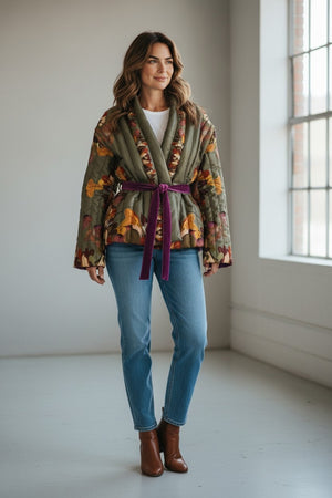 A woman stands indoors near large windows, smiling while wearing the FRANCO FERRARI REVERSIBLE KIMONO JACKET with a botanical floral print, styled with a purple belt, white top, blue jeans, and brown ankle boots.