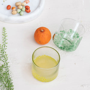 A round tray displays two CREATIVE COOP HAND-BLOWN ROUND CONFETTI DRINKING GLASSES with beverages, decorative stirrers, a green bottle, lemonade bottle, and a small cactus ornament on a tabletop with light linens.