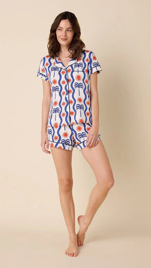 A person in CAT'S PAJAMAS - RED, WHITE AND BREEZE PIMA PAJAMA SHORT SET from CAT'S PAJAMAS stands barefoot sideways on a beige background, glancing back over their shoulder.