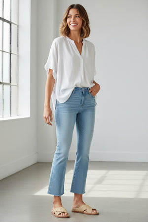 A person wears MERSEA’s Murphy Mini Boot Cut Jean in Sea Spray—light blue, 4-way stretch denim—paired with a white top and beige slide sandals, standing on a white floor before a beige fabric backdrop. Their face is not visible.