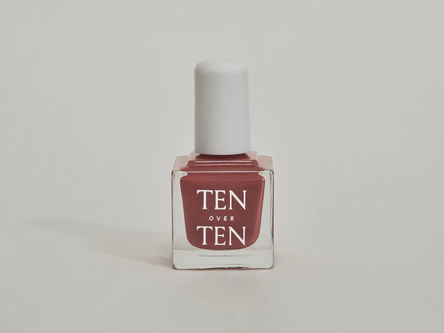 A glass bottle of TENOVERTEN - NAIL POLISH from the brand TEN OVER TEN, featuring a white cap and a dusty pink nail color. The front label displays "TEN OVER TEN" in white uppercase letters. This vegan, eight-free formula ensures both beauty and peace of mind. The background is plain and light-colored.