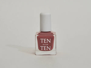 A glass bottle of TENOVERTEN - NAIL POLISH from the brand TEN OVER TEN, featuring a white cap and a dusty pink nail color. The front label displays "TEN OVER TEN" in white uppercase letters. This vegan, eight-free formula ensures both beauty and peace of mind. The background is plain and light-colored.