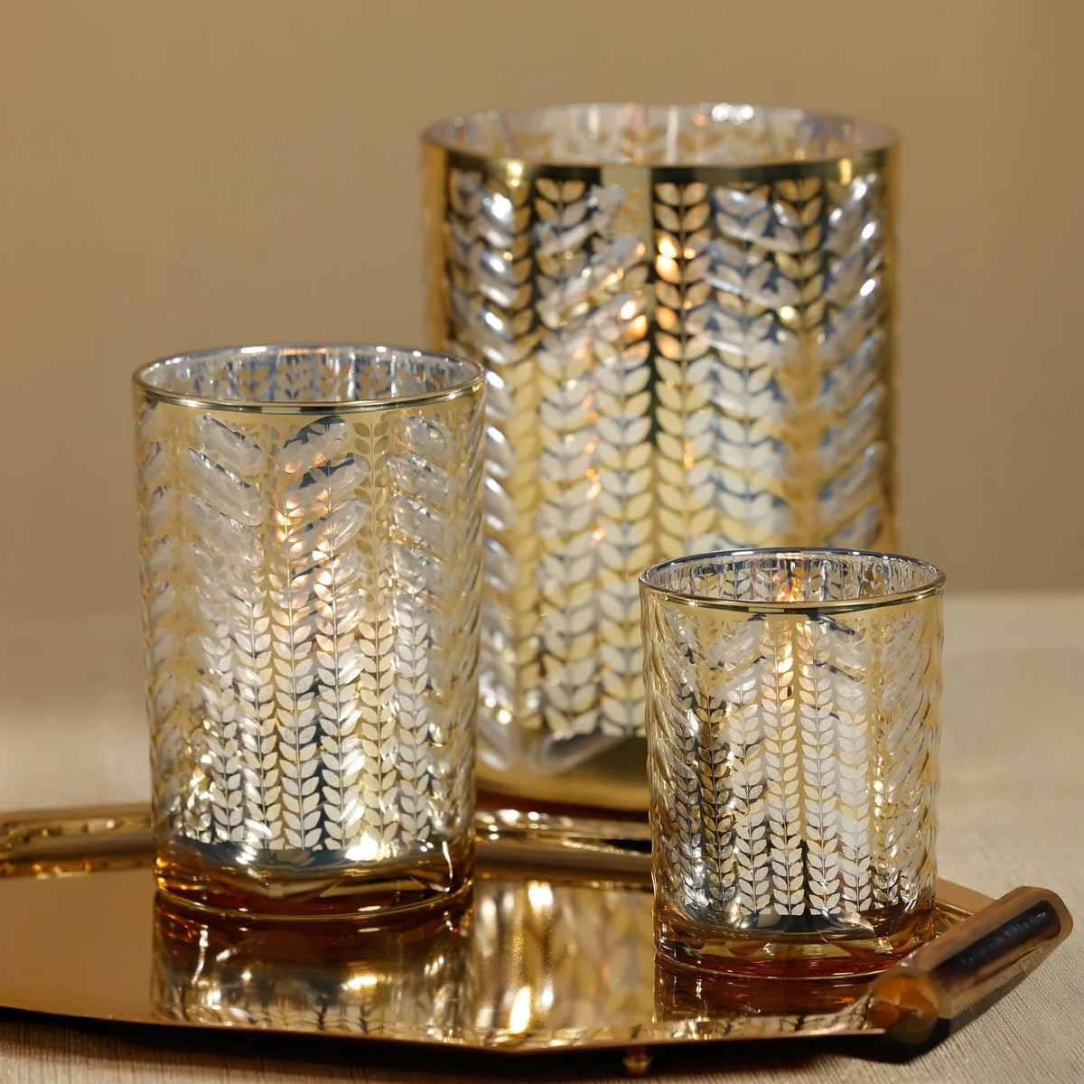 Three 4.5 in x 7 in cylindrical LARGE ETCHED METALLIC HURRICANE 7″ glass candle holders with a metallic gold leaf pattern, arranged on a matching gold serving tray.