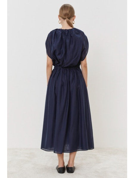 Wearing the ankle-length dark blue MEIMEIJ - OVERLAY DRESS, this person stands against a white backdrop. The dress features gathered sleeves and an elasticized waist, accessorized with black flats and large round earrings, while their hair is neatly pulled back.