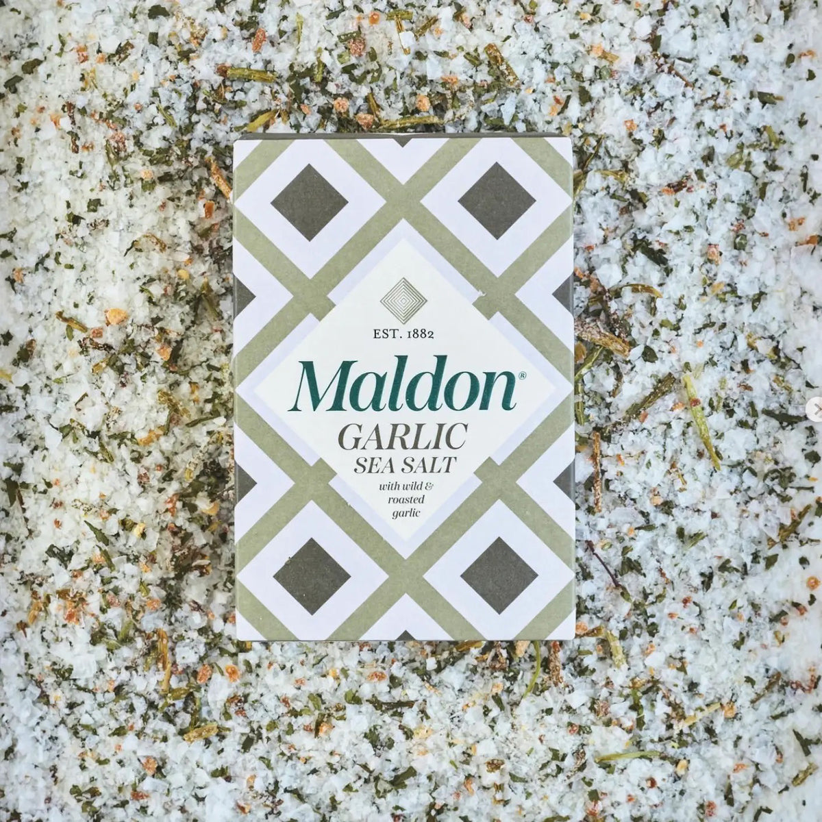 A box of GOURMET FOOD SOLUTIONS&#39; MALDON - GARLIC SEA SALT is placed on a bed of coarse salt with herbs and garlic flakes scattered around. The box, in a green and white geometric design, reads &quot;MALDON - GARLIC SEA SALT&quot; and &quot;with white &amp; roasted garlic,&quot; established 1882—proudly showcasing its English sea salt heritage.