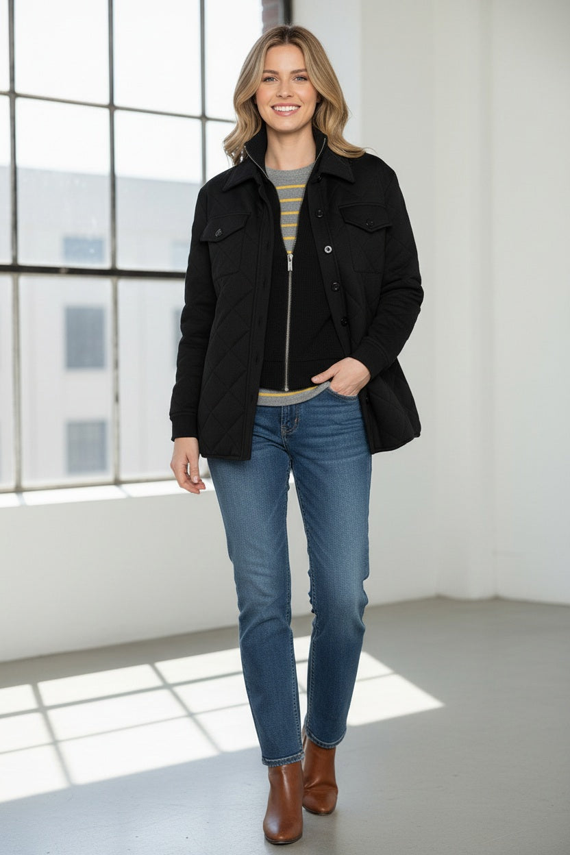 A woman stands indoors by a window, smiling softly as she touches her KINROSS CASHMERE TWO IN ONE QUILTED SHACKET, styled over a striped sweater and blue jeans.