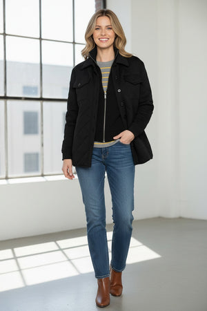 A woman stands indoors by a window, smiling softly as she touches her KINROSS CASHMERE TWO IN ONE QUILTED SHACKET, styled over a striped sweater and blue jeans.
