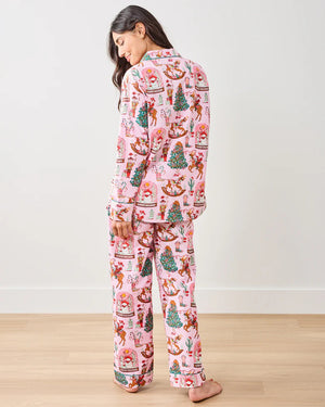A woman sits on the floor against a plain white wall, wearing the PRINTFRESH - COWBOY CHRISTMAS LONG PAJAMA SET by PRINTFRESH, featuring pink fabric with festive Santa, reindeer, wreath prints, and "Howdy Holidays" text.