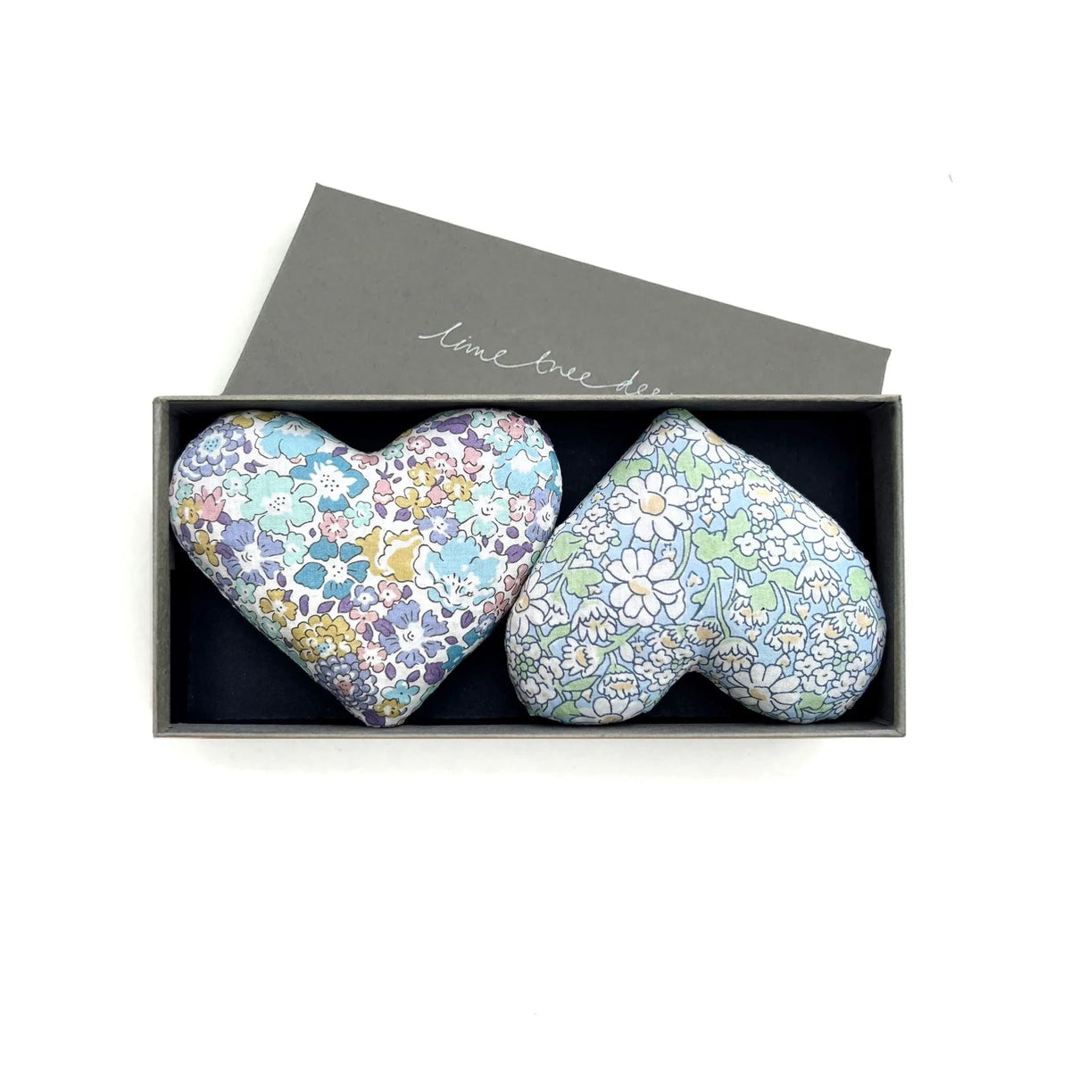 Two colorful heart-shaped sachets from the &#39;HEARTS - SENTIMENTAL SACHET&#39; collection by LIME TREE COLLECTION LTD come in a gray box. Handmade from Liberty Tana Lawn fabric, one sachet features a multicolored floral pattern on a light base, while the other showcases a light blue floral design. The open box lid reveals stylish text on top and releases a hint of lavender.