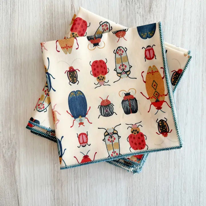 The BEETLE COCKTAIL NAPKIN by DOT AND ARMY features a playful red, blue, and yellow beetle and bug pattern on soft white cotton. Perfect for adding a fun touch to your table setting. Two napkins shown on a light wooden surface.