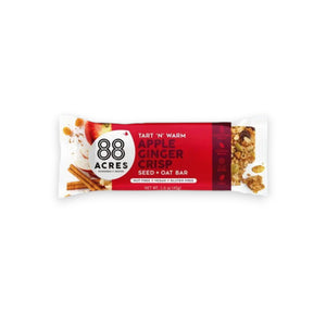 A package of 88 ACRES - APPLE GINGER CRISP SEED + OAT BAR rests on a light wooden surface. The primarily white and red wrapper emphasizes its nut-free, vegan, and gluten-free benefits. The bar is partially visible on the right side.