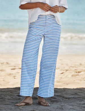 A person stands on a sandy beach facing the ocean, wearing FRANK & EILEEN’s Wexford Wide Leg Linen Pant in Blue Gingham, paired with a crisp white shirt and sandals.