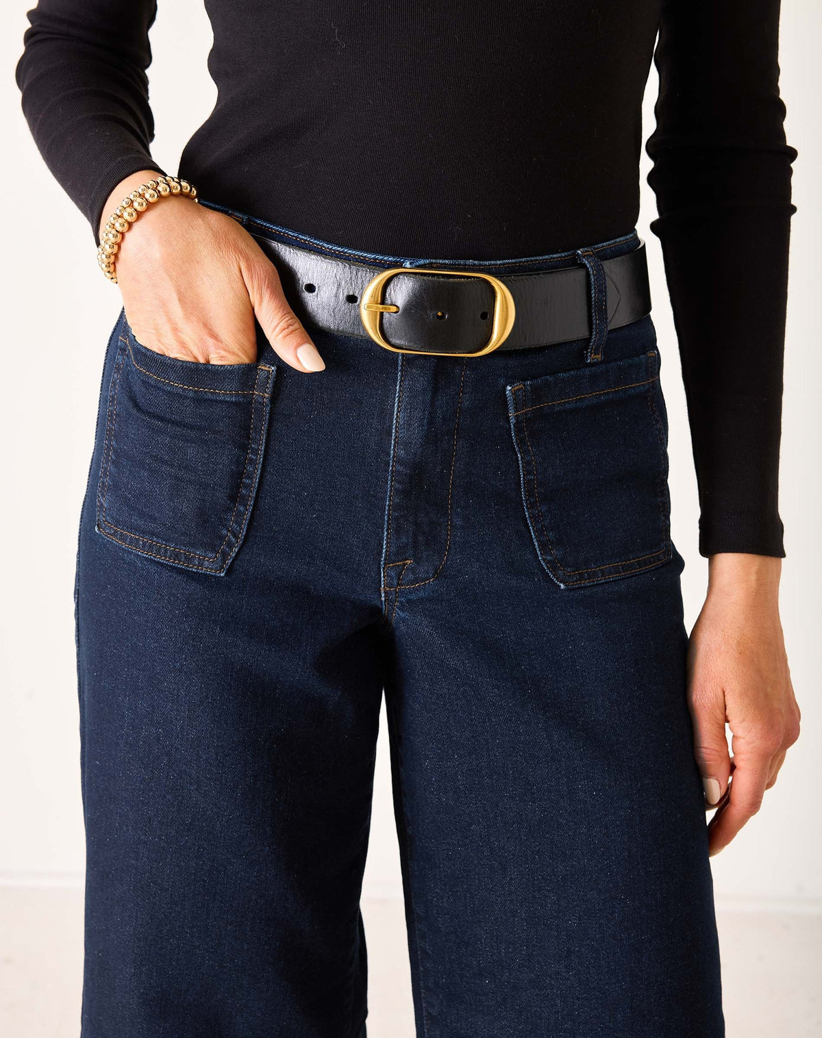 A person wearing MERSEA - SAILOR WIDE LEG JEANS IN RINSE by MERSEA, styled with a black top, black belt, and black heeled sandals, stands with their back to the camera against a plain light background.