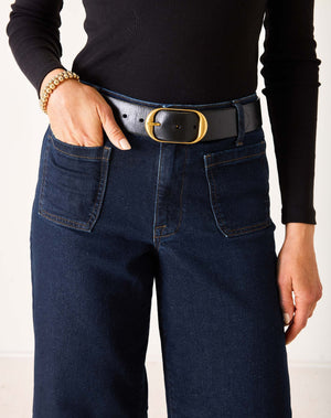A person wearing MERSEA - SAILOR WIDE LEG JEANS IN RINSE by MERSEA, styled with a black top, black belt, and black heeled sandals, stands with their back to the camera against a plain light background.