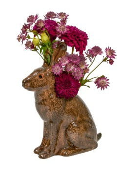 The QUAIL CERAMICS - HARE VASE, LARGE by QUAIL DESIGNS is a hand-painted brown ceramic vase shaped like a hare, displayed with yellow and white flowers and greenery. A single yellow flower lies beside it on a white background.