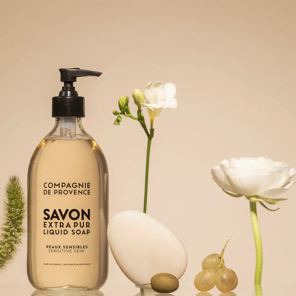 A bottle of CIE LUXE BRANDS COMPAGNIE DE PROVENCE SAVON EXTRA PUR LIQUIDE SENSITIVE SKIN 10OZ is displayed next to a white oval stone, green grapes, a white flower, and green foliage on a beige surface with a neutral background.