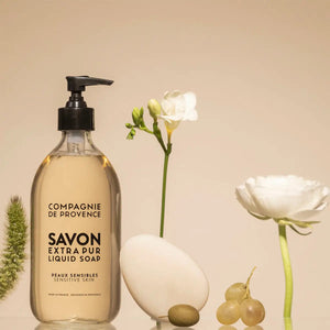 A bottle of CIE LUXE BRANDS COMPAGNIE DE PROVENCE SAVON EXTRA PUR LIQUIDE SENSITIVE SKIN 10OZ is displayed next to a white oval stone, green grapes, a white flower, and green foliage on a beige surface with a neutral background.